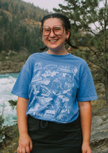 Load image into Gallery viewer, Woman standing by a river wearing a blue t-shirt that has hand-drawn icons for some of Minnesota's top locations
