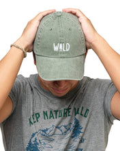 Load image into Gallery viewer, Person wearing a green cap with 'WILD' printed on it and a gray t-shirt with 'KEEP NATURE WILD' printed on it.