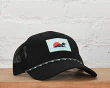 Load image into Gallery viewer, A black trucker hat with a mesh back, resting on a wooden surface against a white brick wall. The front panel features a rectangular patch depicting a red sun over blue water with a black-and-white spotted dog, and a green-and-white patterned rope detail is threaded through the eyelets.