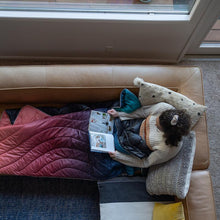 Load image into Gallery viewer, Woman sitting on couch reading with a Rumpl puffy blanket on lap