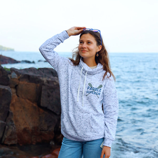 Woman standing by lake with grey cowl neck sweatshirt that says Paddle Superior