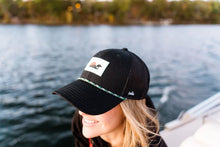 Load image into Gallery viewer, A woman on a boat wearing a black trucker-style cap with a small rectangular logo patch on the front and a green-and-white braided trim along the brim, with water and trees blurred in the background.
