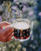 Load image into Gallery viewer, Glass mug with coffee on it. mug is covered in wildflowers and is being held in front of a garden
