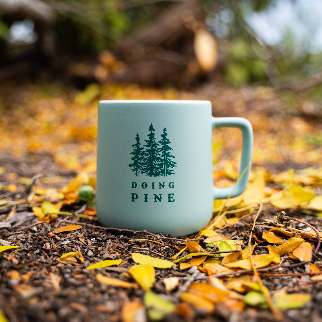 Light blue mug with 'Doing Pine' text and tree design on a forest floor with fallen leaves.