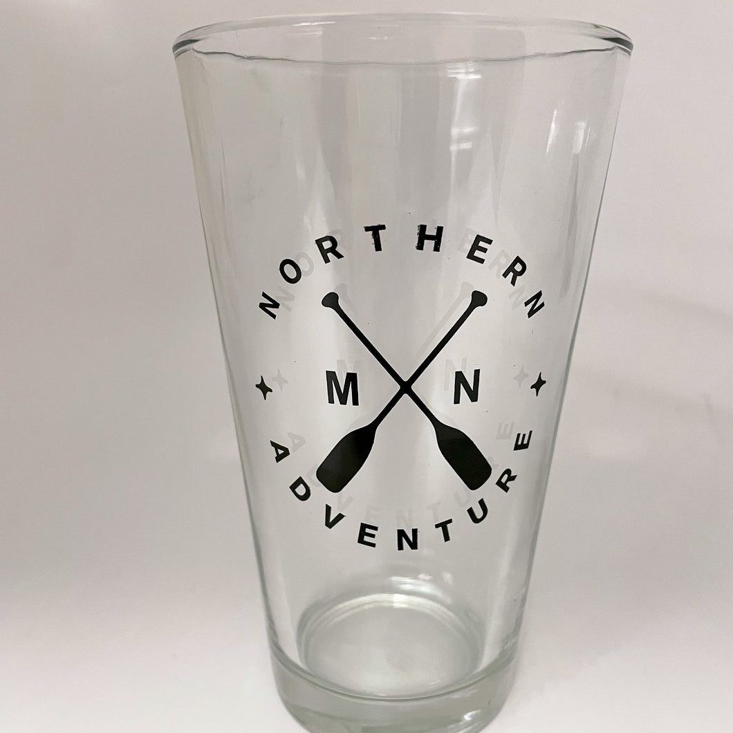 Pint glass with green screen print that says northern adventure MN with cross paddles